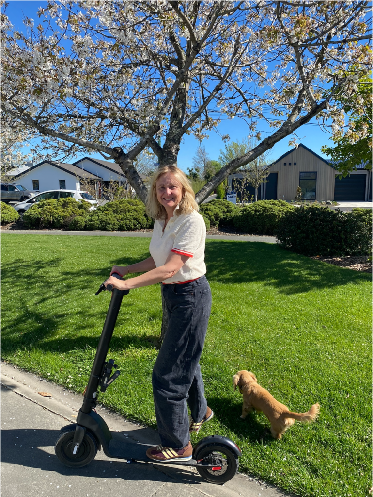 Woman smiling while standing on an electric scooter on a sunny sidewalk, with a small brown dog beside her and blooming trees in the background.