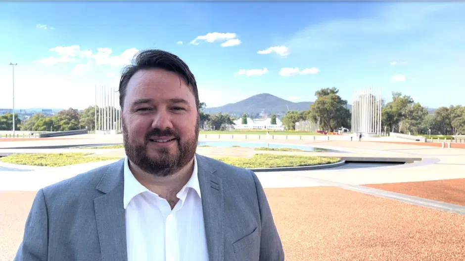 The image features Chris Armstrong, who is in an outdoor setting with a mountainous landscape in the background. He is wearing a suit and standing on a paved area surrounded by greenery and modern structures.